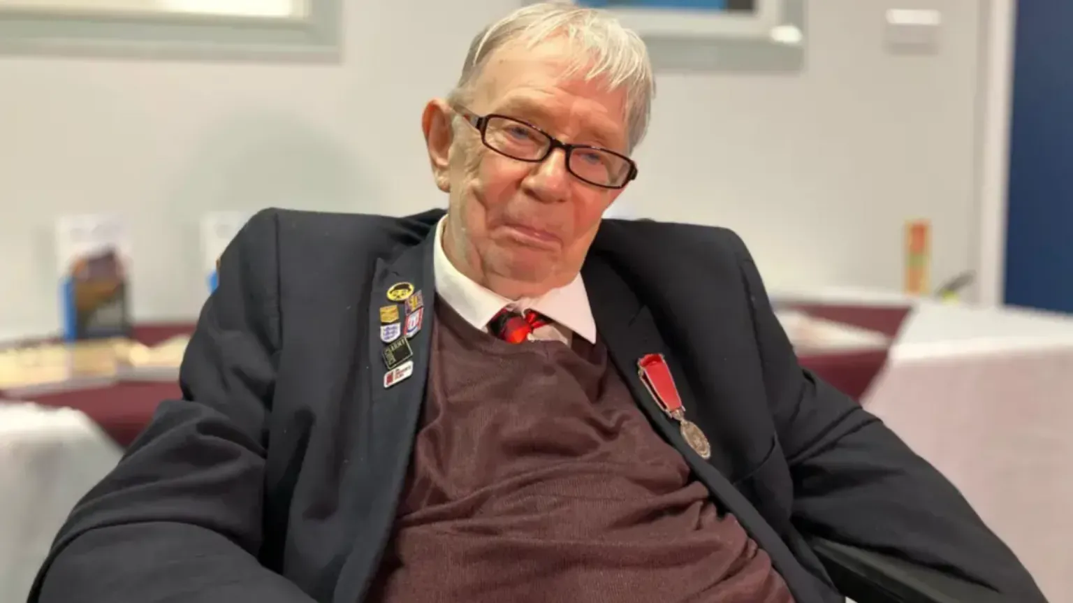 BBC A man wearing a tie, jumper, white shirt and dark jacket is sitting down. The background, including a table and wall, is blurred.