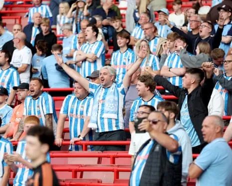 Coventry City fans in striped jerseys cheer with arms raised in stadium seating